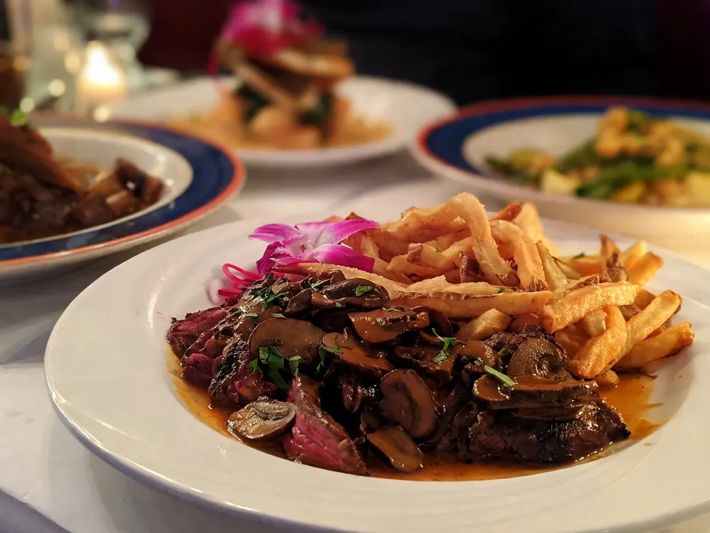 Sliced steak topped with sautéed mushrooms and thin fries on a white plate