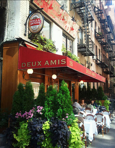 Red awning and sidewalk tables with planters outside a brick corner storefront